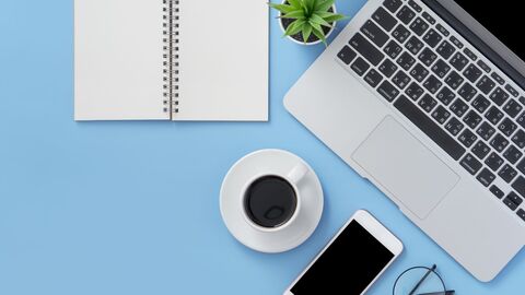 Minimalistisches Bild mit Notizbuch, Laptop, Kaffee, einem Smartphone und einer kleinen grünen Pflanze auf blauem Untergrund.