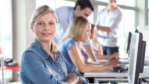 Eine junge Frau sieht lächelnd in die Kamera, im Hintergrund sitzen und stehen Menschen und arbeiten an Computern.
